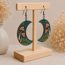 Cargar imagen en el visor de la galería, Enchanted Forest Moon wooden earrings shown at an angle on a light wood stand, highlighting the hand-painted design and natural wood construction.