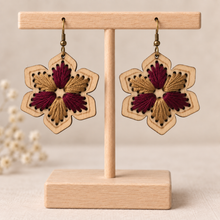 Cargar imagen en el visor de la galería, Hand-embroidered floral wooden earrings in burgundy and warm camel on a light wood stand