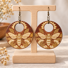 Cargar imagen en el visor de la galería, Sunset Honey Bee engraved wooden earrings with bee and sunflower design on a light wood stand with hypoallergenic bronze-tone hooks