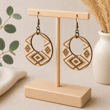 Cargar imagen en el visor de la galería, Angled view of Tribal Spirit engraved wooden earrings on a light wood T-stand, highlighting natural wood grain and the fine laser-engraved geometric lines.