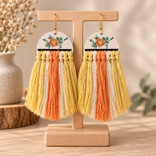 Cargar imagen en el visor de la galería, Sun Whisper handmade wooden earrings with hand-painted floral design and cream, yellow and coral macramé tassels on a light wood stand
