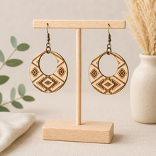 Cargar imagen en el visor de la galería, Tribal Spirit engraved wooden earrings displayed front-facing on a light wood T-stand, showing natural American linden wood grain and bronze-tone hooks.

